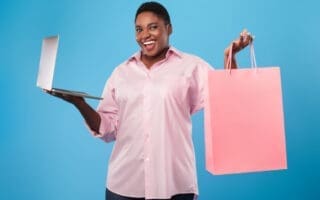 Online shopping. African american woman holding laptop and paper shopper bag standing over blue studio background, smiling to camera. E-commerce, seasonal sales and offers
