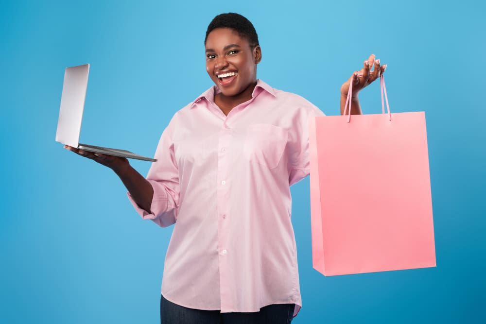 Online shopping. African american woman holding laptop and paper shopper bag standing over blue studio background, smiling to camera. E-commerce, seasonal sales and offers